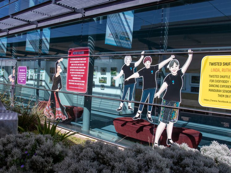 Image of A glass building façade displays large, colourful signs and life-size illustrations of tap dancers. Surrounding plants are visible in the foreground, and reflections show the opposite side of the street.