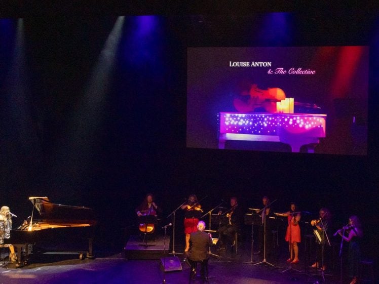 Image of A woman sings beside a grand piano on stage with a string ensemble. A large screen above displays Louise Anton & The Collective with an image of a piano and candles. Stage lighting is purple and blue.