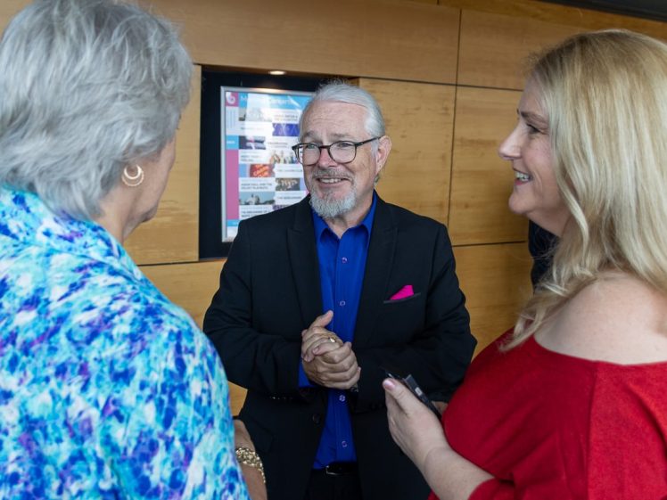Image of Three people stand together indoors, engaged in conversation. An older man in a dark suit and blue shirt smiles at two women, one in a blue patterned blouse and another in a red top, against a wooden wall with a poster in the background.