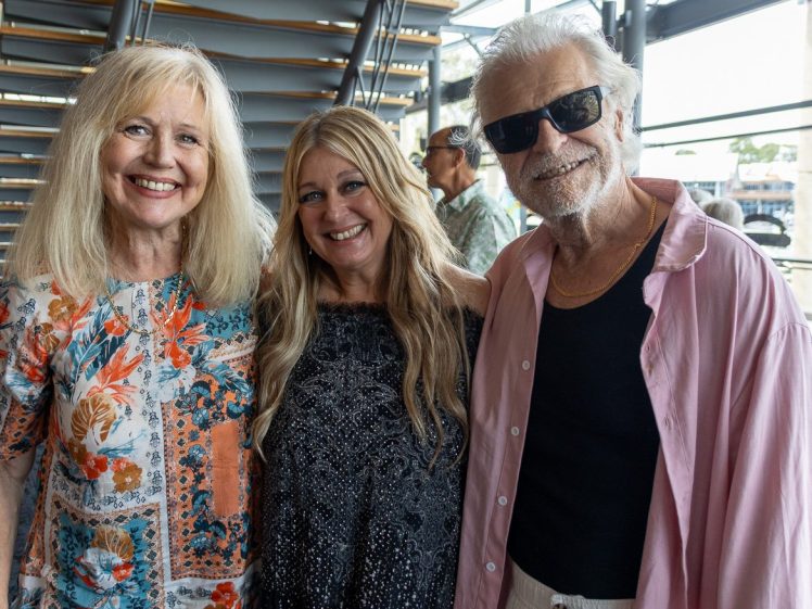 Image of Three smiling adults pose together indoors. The woman on the left wears a colourful floral shirt, the woman in the centre has long blonde hair and a black dress, and the man on the right wears sunglasses, a pink shirt, and a black top.
