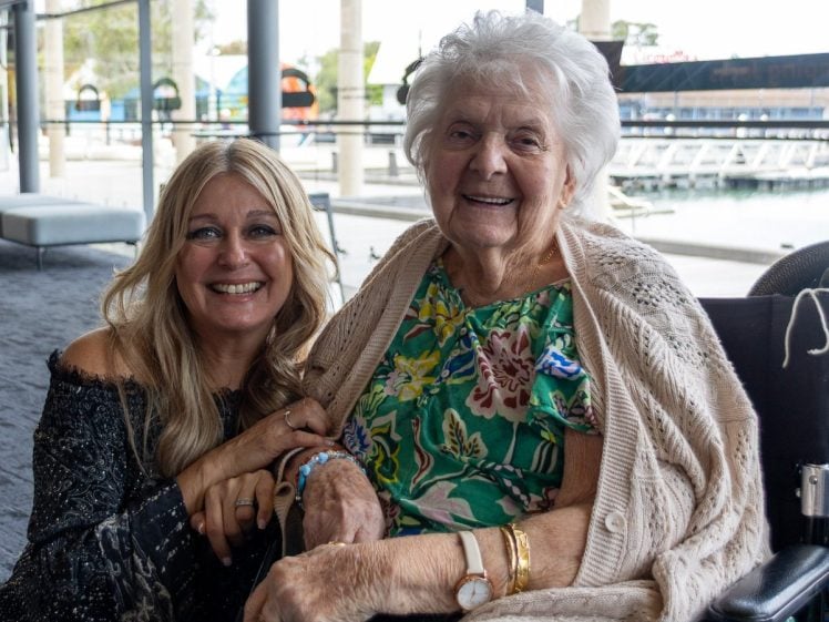 Image of Two women smiling indoors; one is an older woman in a wheelchair wearing a green floral dress and a beige shawl, and the other is a younger woman with long blonde hair, wearing a dark off-the-shoulder top, kneeling beside her.