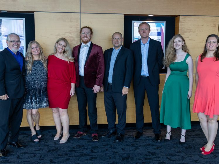 Image of Eight people stand side by side, dressed in formal and semi-formal attire, smiling in front of a wooden wall with framed posters. Three women wear dresses in red, black, and green. Five men wear suits or blazers.