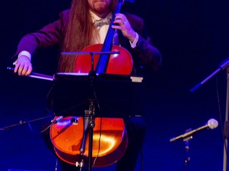 Image of A cellist with long hair and glasses, dressed formally in a suit and bow tie, performs on stage under dramatic lighting, concentrating as he plays his cello with a bow. Sheet music and microphones are visible in front of him.