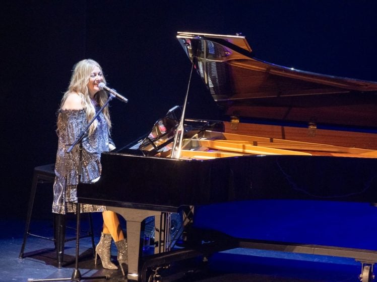 Image of A woman in a sparkly dress and boots sits at a grand piano onstage, singing into a microphone under bright stage lighting.
