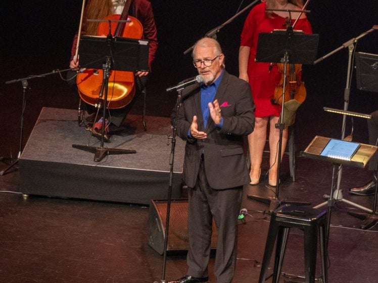 Image of A man in a suit speaks or sings into a microphone on stage, whilst musicians with string instruments sit behind him, including a cellist and a violinist. Music stands and microphones are also visible.