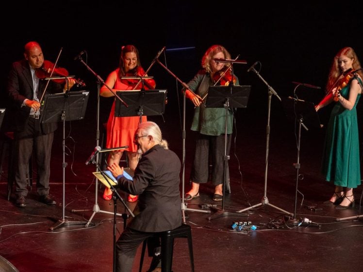 Image of A conductor leads four violinists playing onstage, each standing behind music stands and microphones. The musicians wear colourful dresses or suits, and the stage is dimly lit.