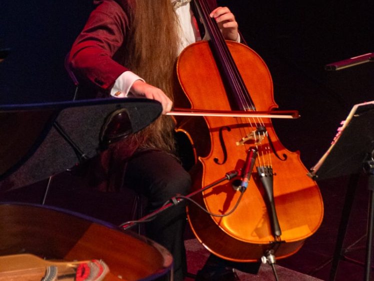 Image of A man with long hair and glasses plays a cello on stage, seated next to a piano and music stand, under warm stage lighting.