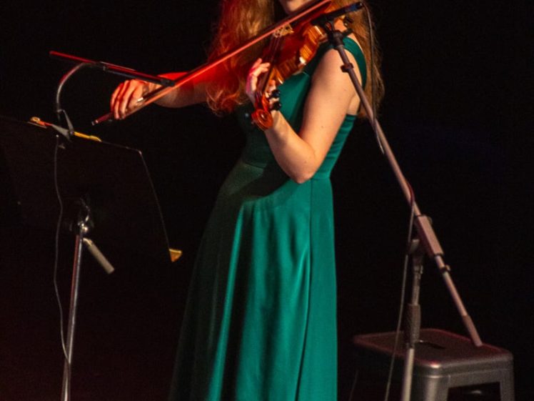 Image of A woman in a green dress stands on stage playing a violin, looking towards the camera with a smile. A music stand and microphone are positioned nearby, and the background is dark.