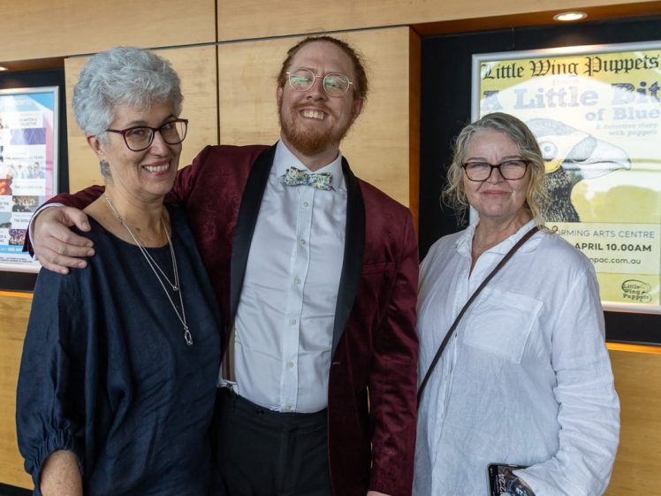 Image of Three adults stand smiling together indoors. The man in the middle wears a maroon jacket and bow tie, with two women on either side. A colourful poster and event boards are visible on the wooden wall behind them.
