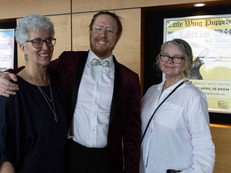 Image of Three people stand together indoors, smiling at the camera. The person in the centre wears a dark red jacket and bow tie, while the two women on either side wear glasses and casual tops. Posters are visible on the wall behind them.