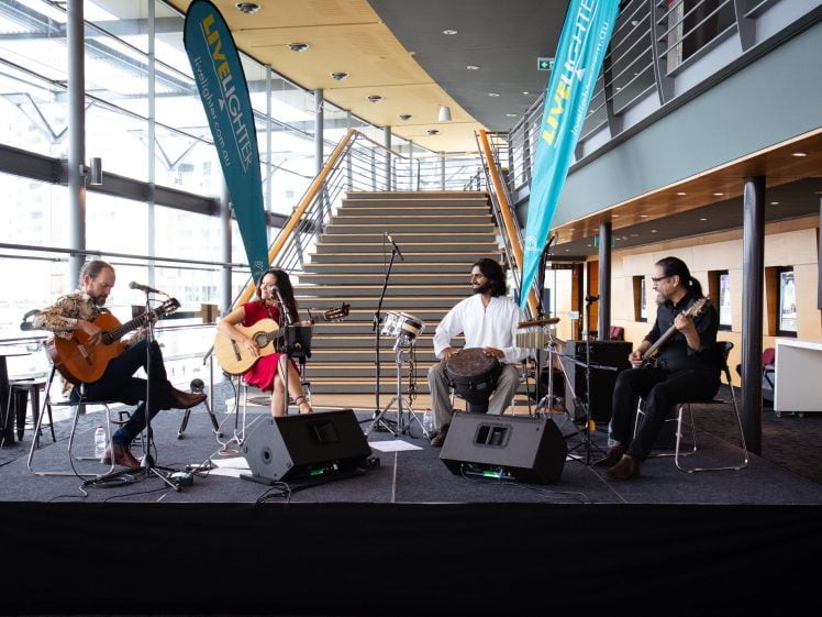Image of Four musicians sit on a stage playing guitar, bass, and drums in a modern indoor venue with large windows and a staircase in the background. Two tall banners and microphones are visible on the stage.