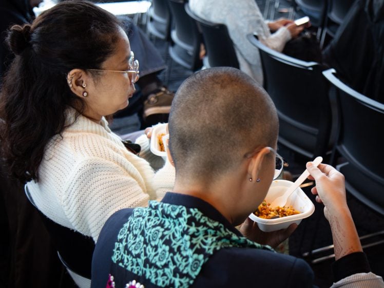 Image of Two people sit side by side in chairs, eating food from white bowls with forks. One wears glasses and a white jumper, the other has short hair and a patterned jacket. Rows of black chairs are visible in the background.