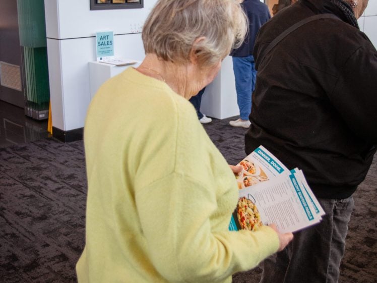 Image of An older woman in a yellow jumper looks at a leaflet or menu whilst standing in an art gallery with several other people. Framed artwork is visible on the walls in the background.