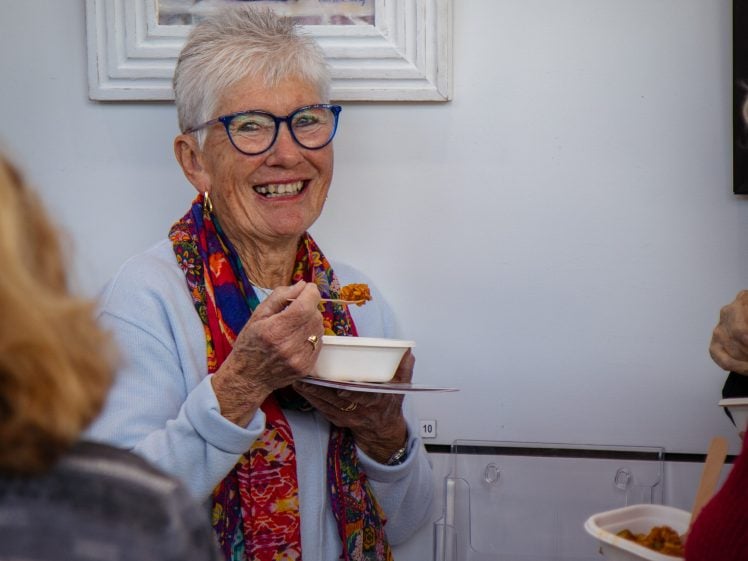 Image of An older woman with short grey hair, blue glasses, and a colourful scarf smiles whilst holding a bowl and spoon at a social gathering. She stands near a white wall with framed art. Other people are partially visible.