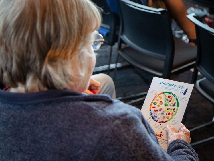 Image of An older person in a blue jumper sits among rows of chairs, holding and reading a colourful healthy eating guide leaflet with a food group diagram.