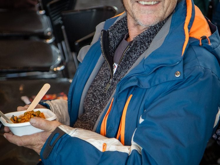 Image of A man wearing a blue and orange jacket sits and smiles whilst holding a bowl of food with a wooden fork. Rows of empty chairs are visible in the background.
