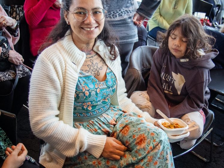 Image of A smiling woman in glasses and a floral dress sits holding a bowl of food, with a young child in a hoodie sitting beside her. Several people are seated in the background, suggesting a community or social gathering.