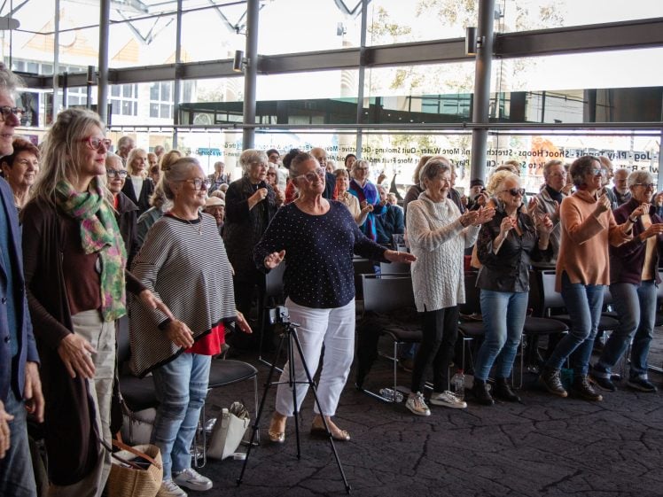 Image of A group of older adults stand and clap enthusiastically at an indoor event, with natural light streaming through large windows behind them. Some people are smiling and interacting, creating a lively and joyful atmosphere.