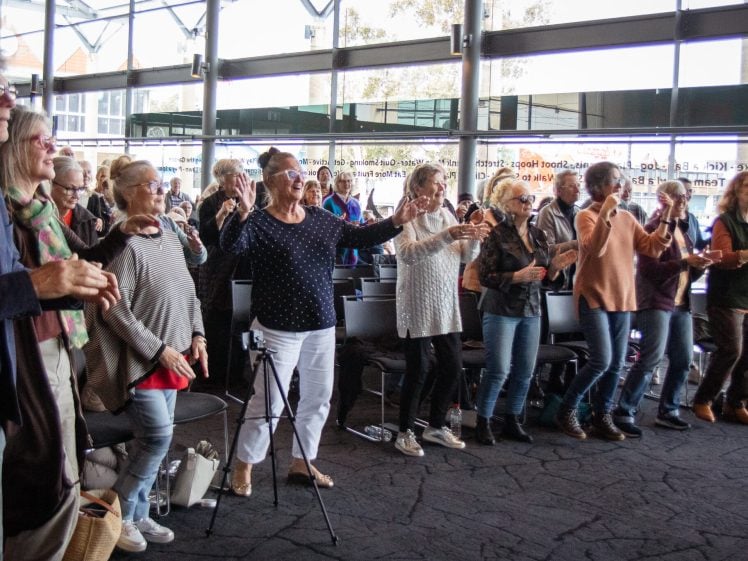 Image of A group of older adults stand indoors in front of rows of chairs, smiling and participating in an activity with their arms raised. Large windows let in daylight; some people are seated, while most are standing and engaging.