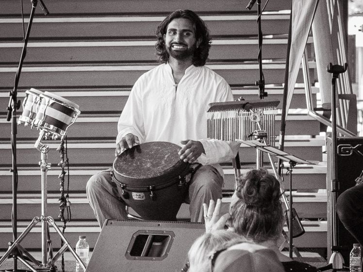 Image of A man with long hair and a beard, wearing a light shirt, smiles while playing a hand drum on stage. Members of the audience watch and gesture in the foreground. Other percussion instruments are nearby.