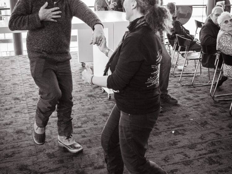 Image of Two people joyfully dance together indoors whilst others sit and watch in the background. The atmosphere is lively, and sunlight streams through large windows. The image is in black and white.