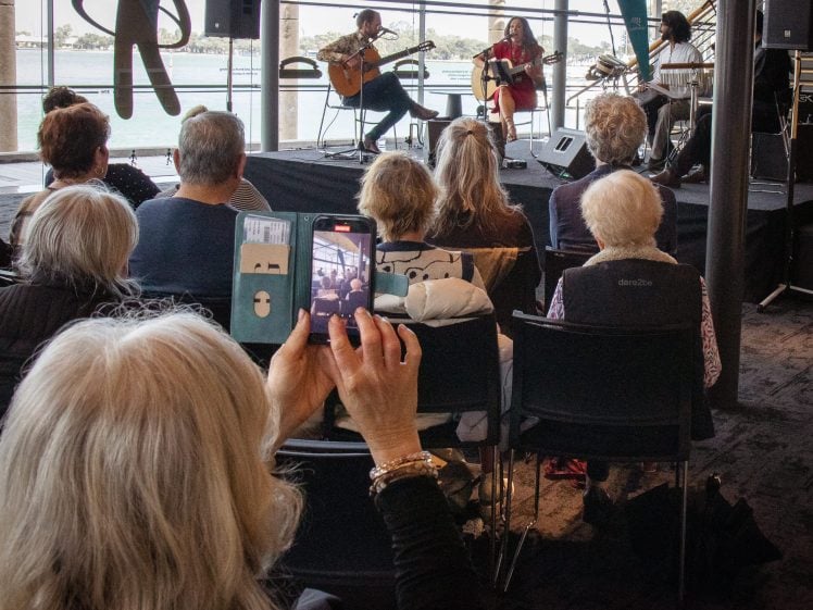 Image of Audience watching a musical performance by three artists on stage near large windows overlooking water, while a woman in the foreground records the scene with her mobile phone.