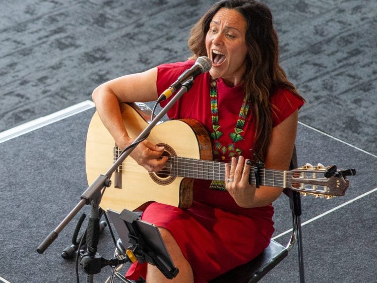 Image of A woman in a red dress sits on a chair, playing an acoustic guitar and singing passionately into a microphone on a stand. She is on a dark carpeted stage with equipment around her.