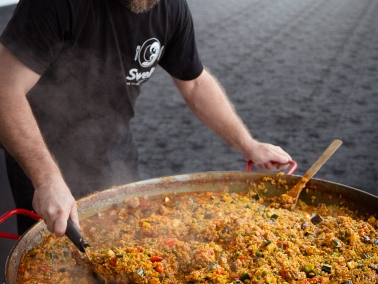 Image of A man with a beard stirs a large pan filled with colourful paella, steaming with rice, vegetables, and meat, at an indoor event.