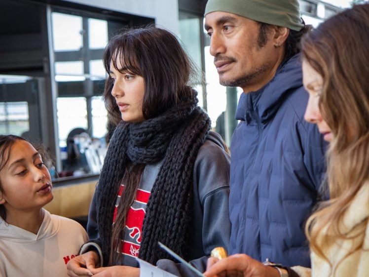 Image of Four people stand indoors. A young girl on the left talks to a woman wearing a black scarf. Next to her, a man in a beanie and blue jacket looks ahead. A woman on the right holds papers and a pen.