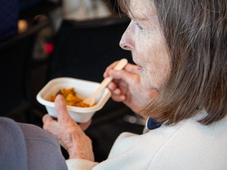 Image of An elderly woman with grey hair, wearing a white jumper, sits and eats food from a small white bowl with a spoon. She is viewed from the side and appears to be at a gathering or event.