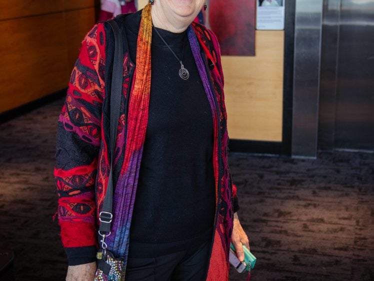 Image of An older woman with short white hair, glasses, and a black hat smiles whilst wearing a colourful red and purple cardigan over a black outfit, standing indoors with a multicoloured handbag and holding spectacles in her hand.
