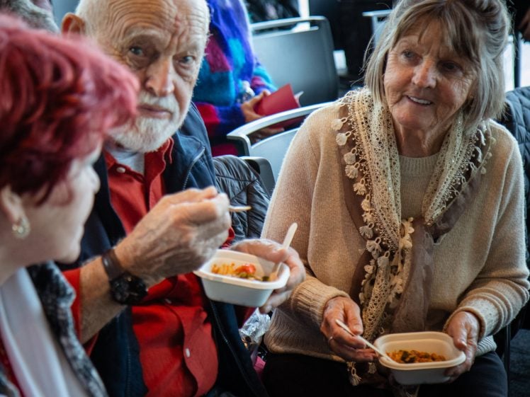 Image of Three older adults sit together, smiling and eating food from bowls with chopsticks, engaged in conversation in what appears to be a social or community setting.