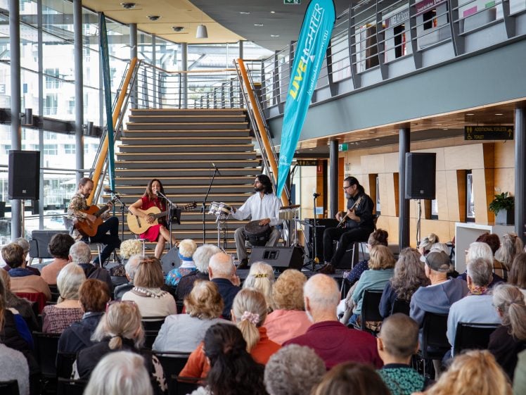 Image of A diverse group of musicians performs on a small indoor stage in front of a seated audience. Large windows and a staircase are in the background, with a blue event banner displayed near the performers.