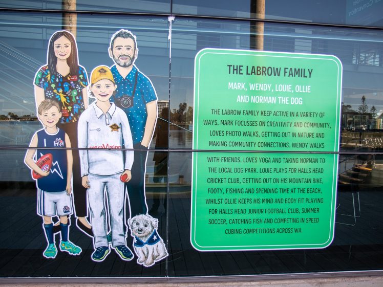 Image of A window display features an illustrated family of four and a dog beside a large green sign introducing the Labrow Family and describing their hobbies, including sport, creativity, dog walks, and outdoor activities.