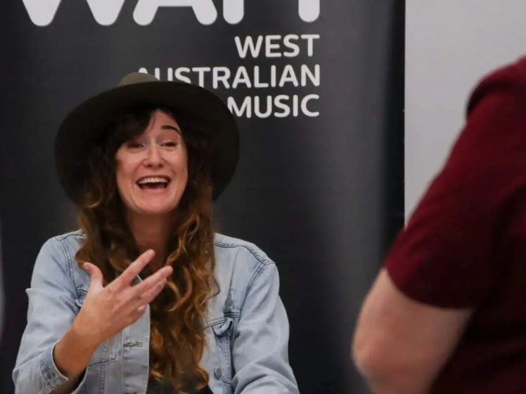Image of A woman with long hair, wearing a wide-brimmed hat and denim jacket, gestures whilst speaking and smiling at someone off-camera. Behind her is a banner reading WAM West Australian Music.