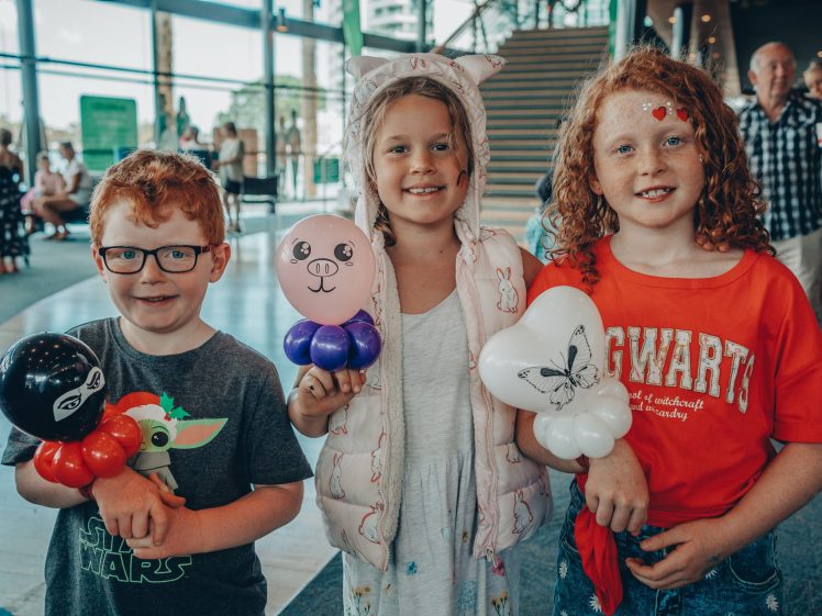 Image of Three smiling children hold balloon animals indoors. The boy on the left wears glasses and a Star Wars shirt, the girl in the centre wears animal ears, and the girl on the right wears a red Hogwarts shirt. Other people are in the background.