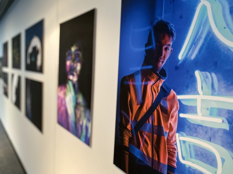 Image of Photographs are displayed on a gallery wall; the closest image shows a person in an orange jacket standing beside bright blue neon lights with Asian characters. Other artworks are blurred in the background.