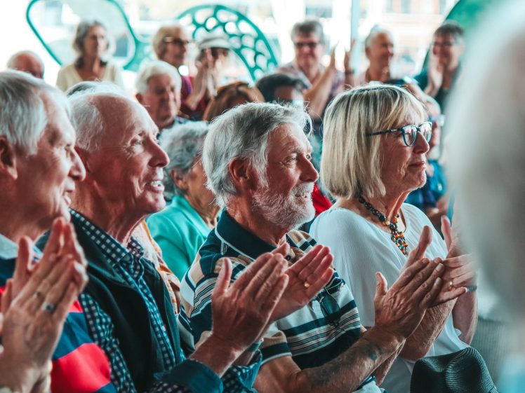 Image of A group of older adults sits closely together, smiling and clapping, appearing engaged and entertained at an indoor event or gathering. The atmosphere is lively and cheerful.