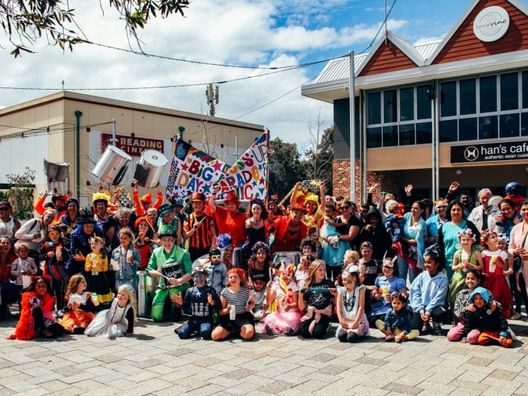 Image of A large group of children and adults, some in colourful costumes, gather outdoors in front of shops for a group photo. The event appears festive, with balloons, banners, and a bright, sunny sky overhead.