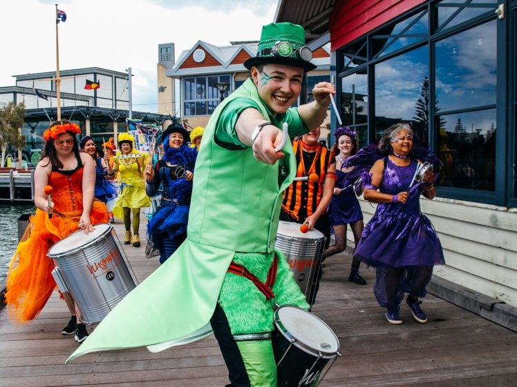 Image of A joyful group of people dressed in colourful costumes plays drums while walking outside. The person in front, wearing bright green and pointing at the camera, leads the lively parade near a waterfront building.