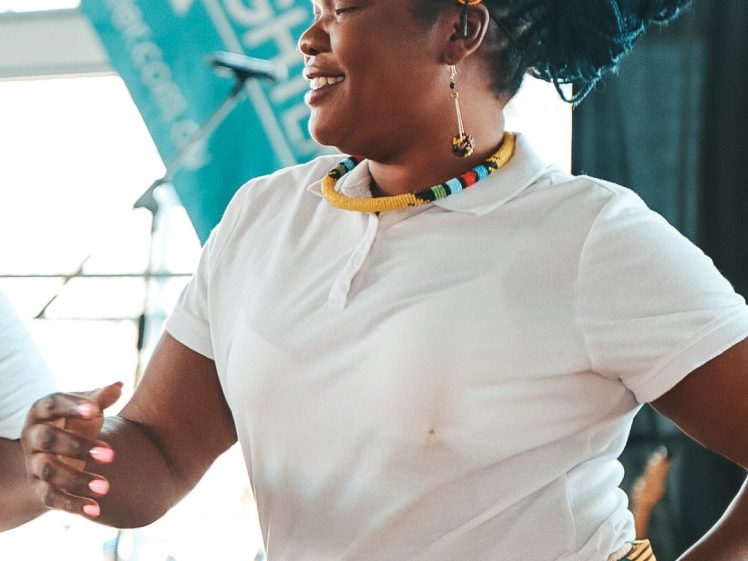 Image of A woman in a white shirt, beaded necklace, and patterned headband and skirt dances and smiles. A turquoise LIVELIGHT banner and blurred background indicate a lively indoor event.