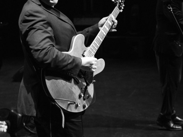 Image of A musician in a suit smiles whilst playing an electric guitar on stage, with a microphone and another person partially visible in the background. The image is in black and white.