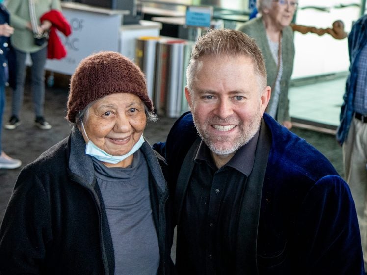 Image of An elderly woman in a brown knitted hat and a younger man in a blue jacket pose together, smiling at the camera in a busy indoor setting with other people in the background.