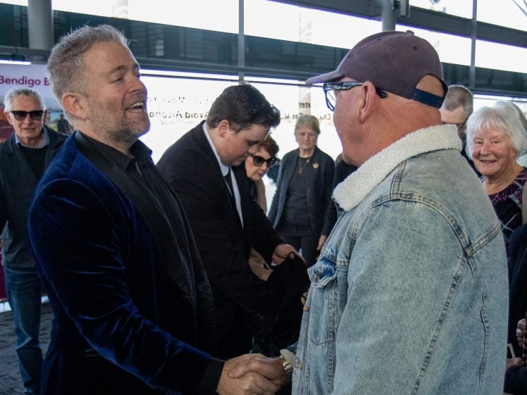 Image of A man in a blue velvet jacket warmly shakes hands with another man in a denim jacket and cap, surrounded by people in a well-lit indoor setting.