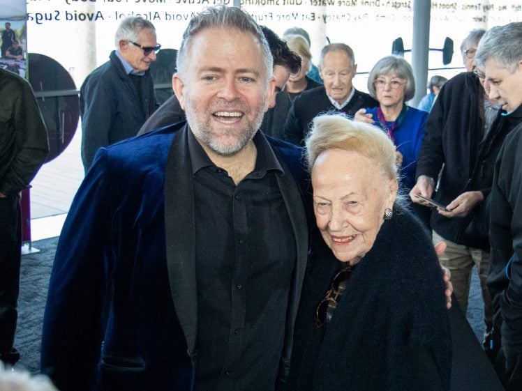 Image of A smiling man in a dark suit stands with an elderly woman in a black outfit at a crowded indoor event. People converse and mingle in the background.