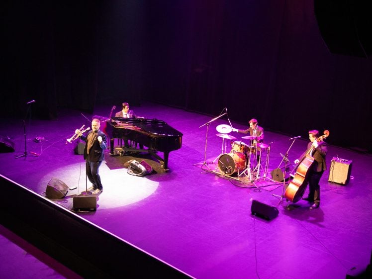 Image of A jazz band performs on a stage under purple lighting, featuring a singer with a trombone, a pianist, a drummer, and a double bass player.