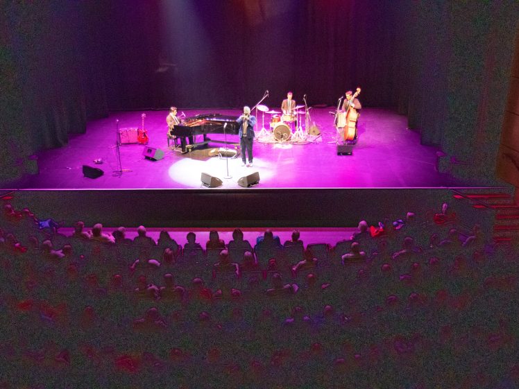 Image of A jazz band performs on stage with a pianist, drummer, double bassist, and a standing vocalist. Purple stage lights illuminate the musicians, whilst the audience watches from the darkened theatre seats.