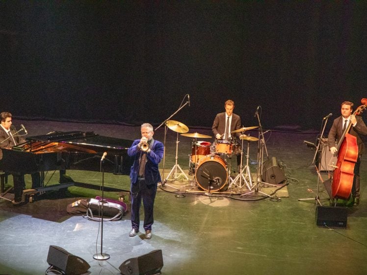 Image of Four musicians perform on stage: one plays the piano, another the trumpet, a third is at the drums, and the fourth plays an upright bass. All are dressed formally, with instruments set up and microphones nearby under stage lighting.