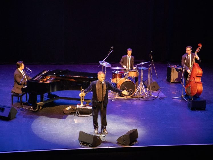 Image of A jazz band performs on stage; a man stands singing at the centre with a trumpet, while musicians play piano, drums, and double bass behind him under stage lighting.