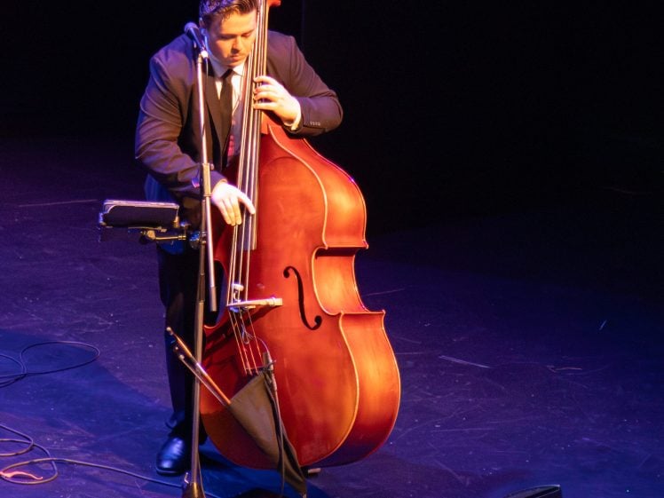 Image of A musician in a suit plays a double bass on stage under bright lighting, surrounded by microphones and audio equipment, with a dark background.